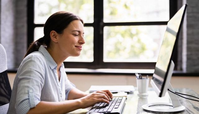 Smiling young woman typing at modern glass desk in bright home office with desktop monitor