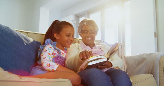 An African American girl and a senior woman are sharing a moment reading a book together on a couch, with copy space. Their bonding over literature highlights the importance of family connections and the transfer of knowledge between generations.