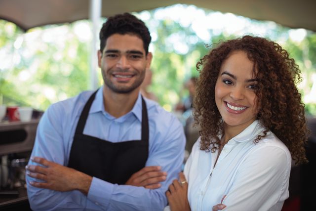 This image shows a smiling waiter and waitress standing with their arms crossed at a counter in a restaurant. They are dressed in professional attire, with the waiter wearing an apron. The background features a bright, blurred outdoor setting, suggesting a pleasant and welcoming atmosphere. This image can be used for promoting restaurant services, hospitality industry, teamwork, and customer service training materials.
