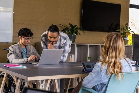 Collaborating diverse team around laptop with wheelchair inclusion in modern daylight office