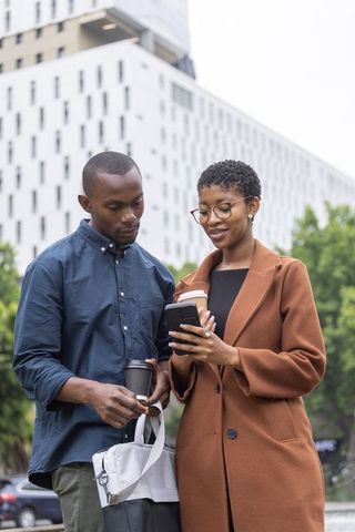 African professionals checking smartphone while holding coffee on urban sidewalk