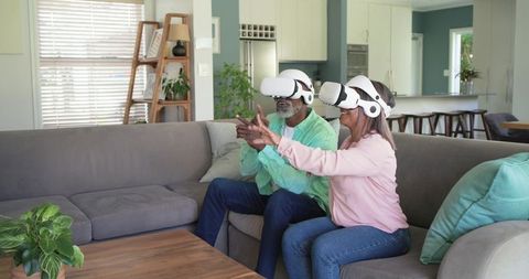 Mature couple wearing VR headsets on gray sectional in modern living room