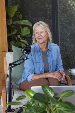 Mature woman hosting podcast in cozy home studio surrounded by greenery and laptop