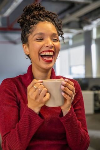 Laughing young african american woman holding mug in modern office wearing red sweater