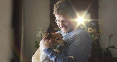 Midlife man cradling small dog in cozy sunlit home, tender canine bond and warmth