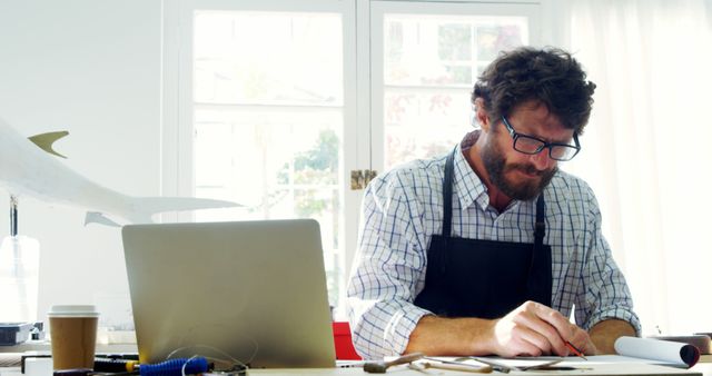 Mature man wearing glasses and a checked shirt engrossed in his work at an office desk. He is surrounded by tools and a laptop, suggesting a creative or technical occupation. Suitable for workplace imagery content, creative professions, and casual office environments.