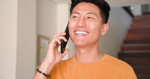 A cheerful Asian man is holding a smartphone to his ear while standing near a staircase. His friendly expression and casual orange t-shirt convey an approachable and modern lifestyle. The warm tones of the interior add to the inviting atmosphere. This image is ideal for advertisements or articles focusing on modern communication, young professionals, or lifestyle content.