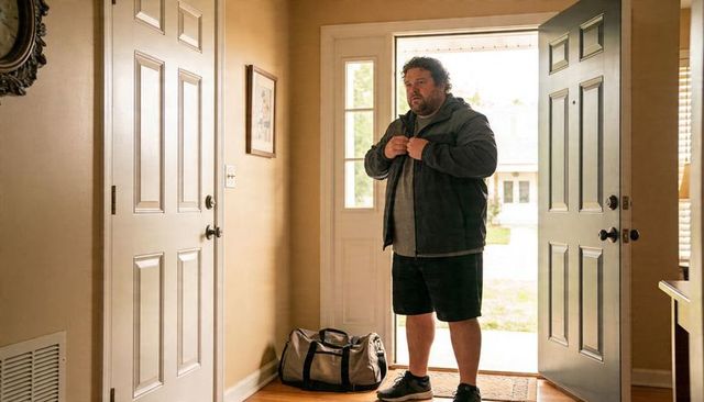 Man buttoning jacket in sunlit suburban entryway with duffel bag and open door
