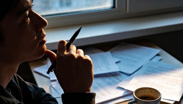 Pensive young man holding pen, gazing out window over sunlit desk with papers and coffee
