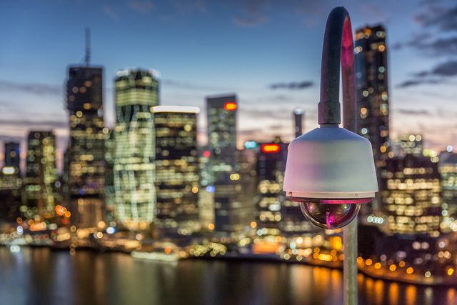 This image features a surveillance camera mounted with a blurred modern cityscape in the background at dusk. Suited for security system advertisements, urban technology presentations, safety promotion materials, and articles about privacy and safety regulations in metropolitan areas.