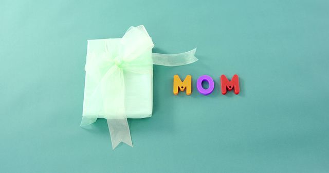 A gift wrapped in pastel paper with a delicate bow is placed next to colorful letters spelling MOM on a soft green background, with copy space. It suggests a celebration of motherhood, for Mother's Day or a birthday, with room for a heartfelt message.