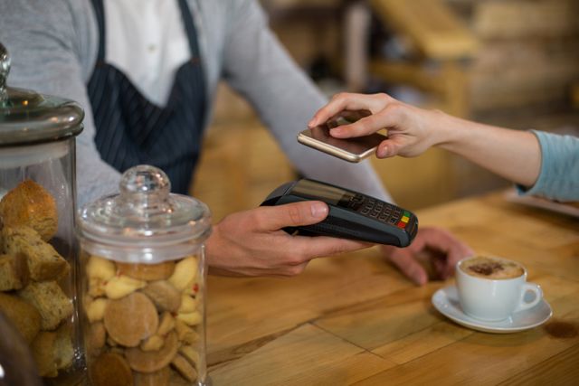 Woman using smartphone for contactless payment in a cafe. Barista holding payment terminal. Ideal for illustrating modern payment methods, technology in retail, and convenience in small businesses.