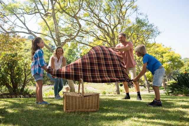 Family enjoying a sunny day in the park, spreading a picnic blanket on the grass. Ideal for concepts related to family bonding, outdoor activities, leisure time, and summer fun. Perfect for advertisements, blogs, and articles about family life, outdoor recreation, and healthy living.