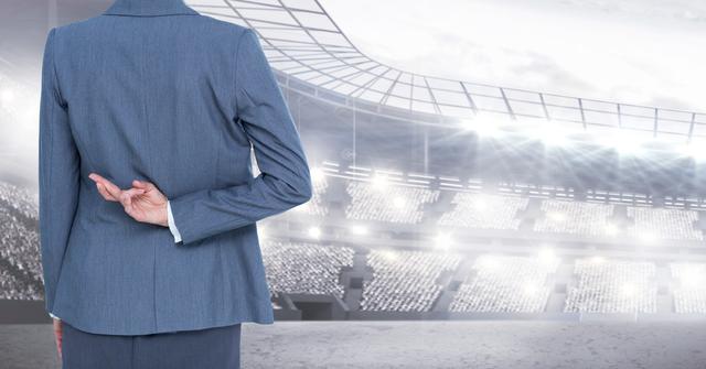 Businesswoman in a suit crossing fingers behind her back while standing in a stadium. This image can be used to represent themes of deception, hope, strategy, and professional competition. Ideal for articles or presentations on business ethics, sports management, or corporate strategy.