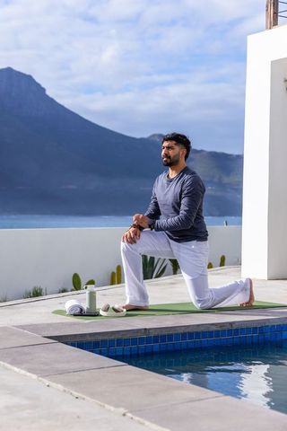 Indian man practicing kneeling lunge yoga poolside on terrace with sea and mountain view