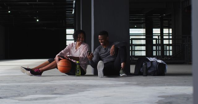 Young couple sitting together with basketball and water bottles, relaxing and smiling after a game. The urban indoor court setting adds a modern and casual vibe, suitable for promoting fitness and active lifestyle, workout programs, recreational sports, and urban living themes.