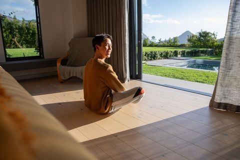Asian man sitting on light wood floor by open sliding door smiling at sunlit backyard pool