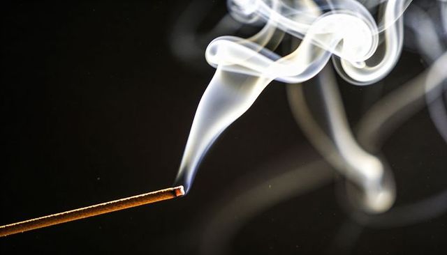 Glowing incense stick emitting curling smoke plumes against dark studio backdrop