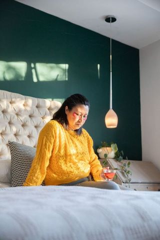 Sunlit senior woman sitting on bed wearing mustard sweater holding pink drink in cozy bedroom