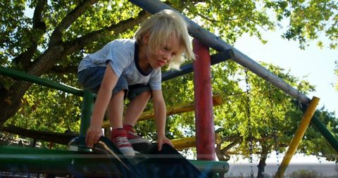 Crouching blond boy gripping slide edge on colorful playground, climbing and playing