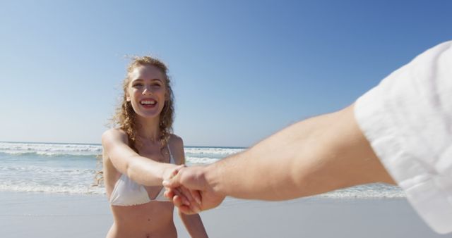 A young Caucasian woman in a bikini smiles as she holds someone's hand on a sunny beach, with copy space. Her joyful expression and the clear blue sky create a carefree, summer vibe.