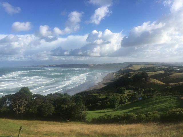 Sweeping view of a coastal landscape featuring rolling waves, expansive beach, and lush green hills beneath a partly cloudy blue sky. Ideal for use in travel brochures, outdoor adventure advertisements, eco-tourism promotions, or as a stunning backdrop in presentations emphasizing natural beauty and scenic tranquility.
