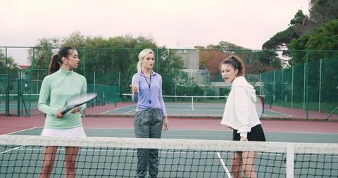 Female tennis coach giving instruction to two young players on outdoor court with rackets