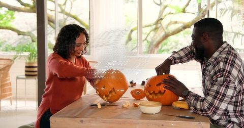 Couple carving jack-o'-lanterns at sunlit wooden table, cozy autumn diy together