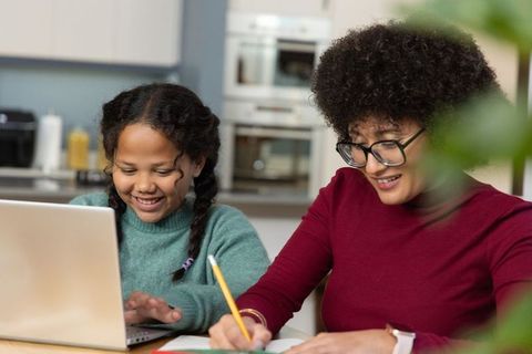 Female guardian and child collaborating on homework at kitchen table with laptop