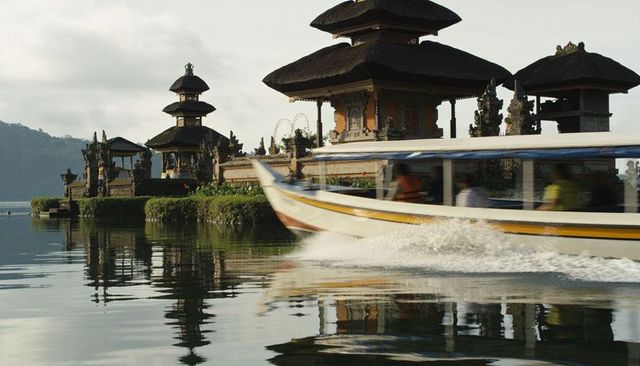 Speeding yellow-striped boat passing balinese lakeside temple leaving foamy wake and reflections