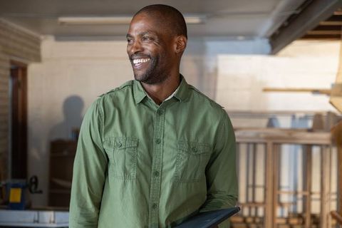 Mature african american craftsman smiling holding tablet in modern carpentry workshop