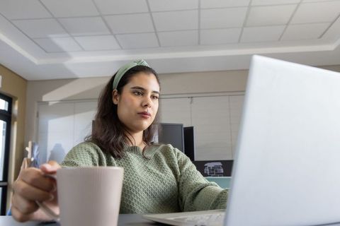 Young asian woman typing on laptop while holding mug in modern sunlit office