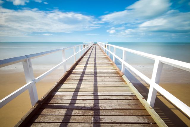Wooden plank walkway extending far out into calm blue sea, framed by white railings under clear blue sky with scattered white clouds. Suitable for advertisements promoting vacation destinations, peaceful retreats, or oceanfront properties. Ideal for use in travel brochures, tourism websites, and lifestyle magazines.