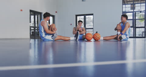 Three young basketball players in sports uniforms sit on an indoor court, engaging in stretching exercises while sharing a moment of laughter. This scene highlights teamwork, camaraderie, and the balance between discipline and enjoyment in sports routines. Ideal for showcasing teamwork, sports training environments, or fitness-focused content.