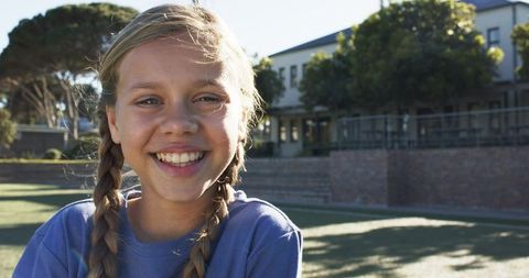 Smiling schoolgirl with braided hair on sunlit campus lawn, casual blue crewneck portrait