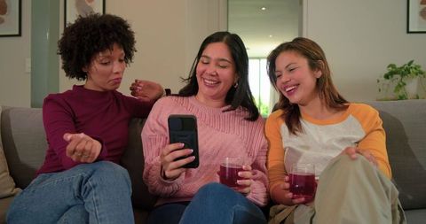 Multicultural women laughing and sharing phone on sofa while drinking wine and relaxing
