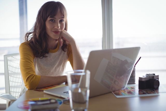 Young businesswoman sitting at a desk in a creative office, working on a laptop. Ideal for illustrating modern work environments, remote work, freelance lifestyle, and professional women in the creative industry.