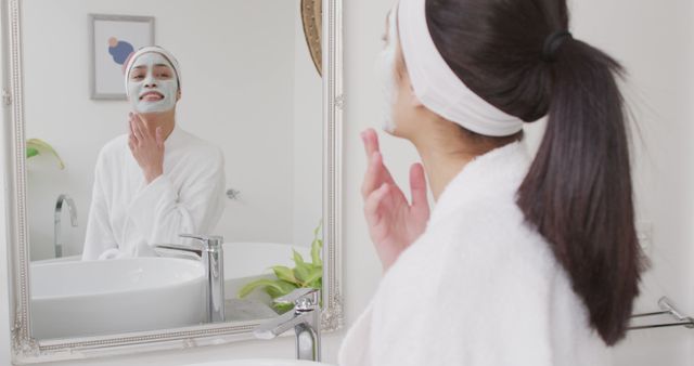 A biracial woman is applying cream on her face in a modern bathroom. She is engaging in a beauty routine focused on skincare and moisturizing. This photo is versatile for many applications including articles on self-care, beauty and skincare products, personal hygiene, or wellness blogs. It emphasizes daily routine practices and healthy skin care habits, perfect for promoting skincare brands or self-care tips.