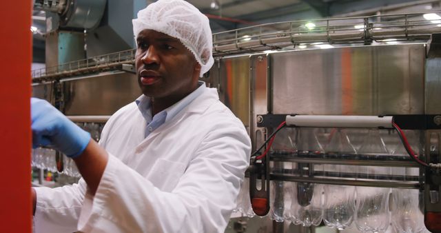 Factory worker wearing white lab coat, gloves, and hairnet inspecting machinery along production line. Ideal for use in articles and advertisements about industrial manufacturing, quality control, health and safety standards in food processing or beverage production environments.