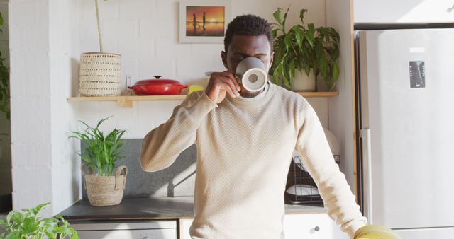 Happy african american man standing in kitchen, drinking coffee. Spending quality time at home alone.