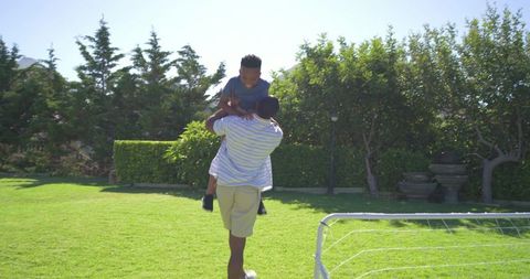 African american father carrying teen son across sunny backyard near soccer goal