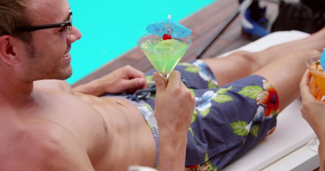 Man lounging beside swimming pool, enjoying a green tropical cocktail with decorative umbrella. Ideal for marketing summer vacations, leisure activities, poolside relaxation, resort advertising, and travel promotions.