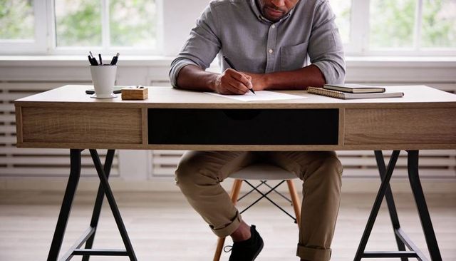 Focused man writing at minimalist wooden desk in bright home workspace with notebook