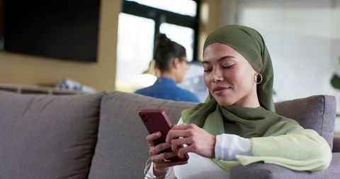 Muslim woman wearing green hijab using smartphone on couch while flatmate working remotely