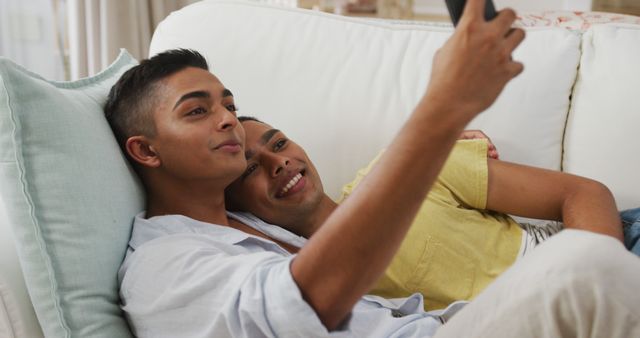 Smiling biracial gay male couple hugging lying on sofa taking selfie. staying at home in isolation during quarantine lockdown.