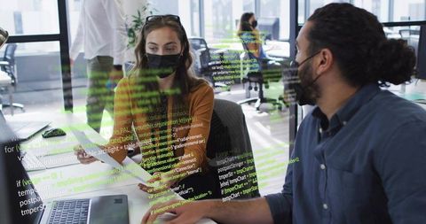 Masked developers collaborating over laptop with code overlay in modern open office