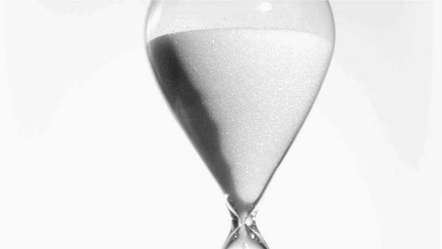 The image showcases an elegant hourglass as sand flows slowly marking the passage of time. Its minimalist design against a white background creates a sense of serenity and focus. This representation can be used in concepts relating to solutions for time management, processes, waiting, or highlighting timeless beauty or sustainability topics.