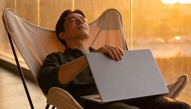 Relaxing asian man reclining with closed laptop in sunlit modern lounge