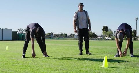Coach overseeing athletes performing warm up stretches between cones on sunny sports field