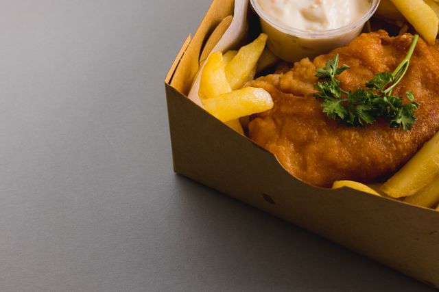 Close-up view of fried seafood and golden french fries in a cardboard takeout box, accompanied by a small container of sauce and garnished with parsley. Ideal for use in food blogs, restaurant menus, fast food advertisements, and culinary websites.